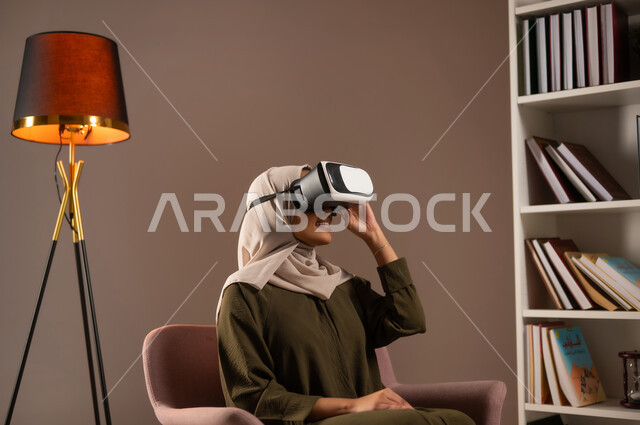 التفاعل والاندماج ومحاكاة العالم الخيالي, الاستمتاع بمشاهدة أفلام الواقع الافتراضي, بورتريه لشابة عربية خليجية سعودية ترتدي العباءة الملونة ونظارة في آر ثلاثية الأبعاد تجلس على الكرسي في المكتبة وتنظر إلى شيء ما بتركيز, الترفيه والتسلية في وقت الفراغ, خلفية بيج