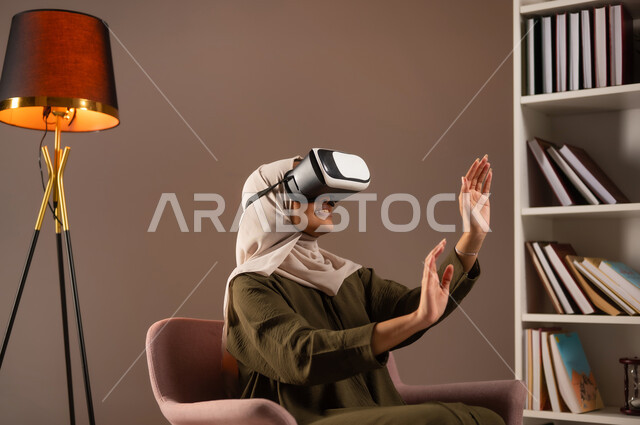 الترفيه والتسلية في وقت الفراغ, الاستمتاع بمشاهدة أفلام الواقع الافتراضي, بورتريه لشابة عربية خليجية سعودية ترتدي العباءة الملونة ونظارة في آر ثلاثية الأبعاد ترفع يديها لمحاولة لمس شيء ما في المكتبة, التفاعل والاندماج ومحاكاة العالم الخيالي, خلفية بيج