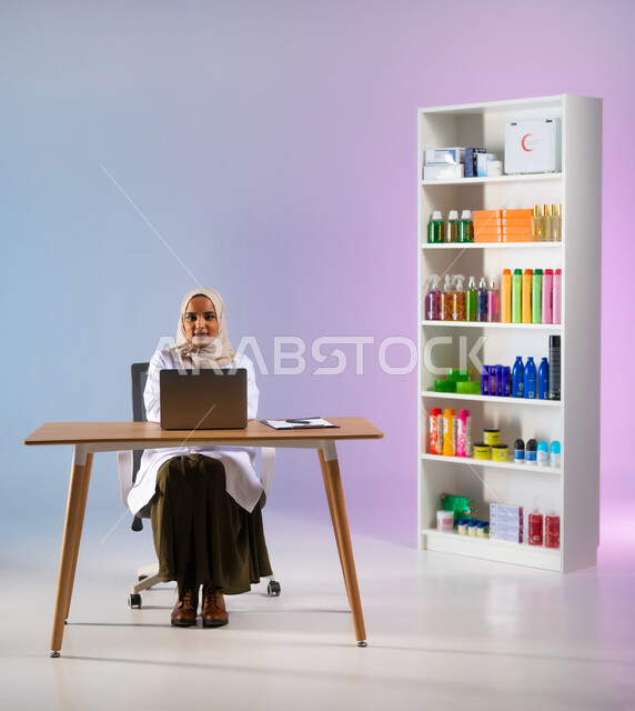 البحث الطبي عن الوصفات العلاجية عبر الاونلاين, مفهوم الصيدلة واستخدام المنتجات الصحية, التسويق والترويج لمتسحضرات العناية بالبشرة, بورتريه لطبيبة صيدلانية عربية خليجية سعودية ترتدي المعطف الطبي الأبيض تقوم بالعمل على الحاسب المحمول, خلفية ملونة