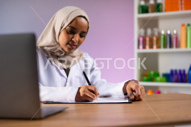استخدام الأجهزة والتقنيات الحديثة والمتطورة, الادوية والمستلزمات الطبية، مفهوم الصيدلة والرعاية الصحية، التشييك على قائمة الأدوية, بورتريه مقرب لطبيبة صيدلانية عربية خليجية سعودية محجبة ترتدي المعطف الطبي الأبيض تقوم بكتابة الوصفات العلاجية, خلفية ملونة