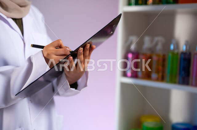 مفهوم الاهتمام بالجسد, عرض المنتجات الطبية في الصيدليات, التسويق للعطور والمستحضرات النسائية, بورتريه مقرب ليد طبيبة صيدلانية عربية خليجية سعودية محجبة مبتسمة ترتدي المعطف الطبي الأبيض تقوم بتدوين وتسجيل مخزون الأدوية والمواد الطبية, خلفية ملونة