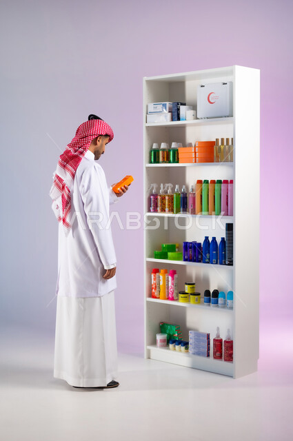 مفهوم الاهتمام بالجسد, قراءة صلاحية المنتج ومكوناته, الإعلان والتسويق لمنتجات صحية، العناية بالشعر والنظافة الشخصية, بورتريه من الجانب لطبيب صيدلاني عربي خليجي سعودي يرتدي المعطف الطبي الأبيض يحمل بيده شامبو الشعر في الصيدلية, صورة الجسم بالكامل, خلفية ملونة
