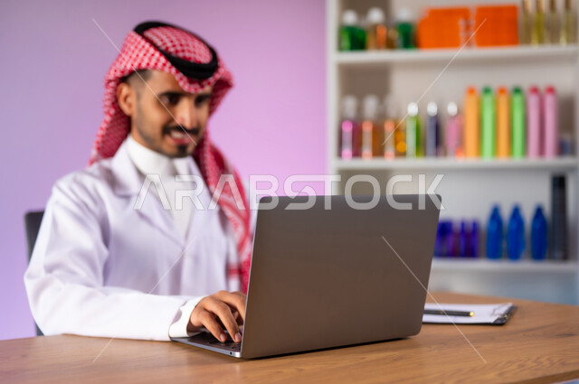البحث الطبي عن الوصفات العلاجية عبر الاونلاين, مفهوم الصيدلة واستخدام المنتجات الصحية, التسويق والترويج لمتسحضرات العناية بالبشرة, بورتريه مقرب لطبيب صيدلاني عربي خليجي سعودي مبتسم يرتدي المعطف الطبي الأبيض يقوم بالعمل على جهاز الحاسب المحمول, خلفية ملونة