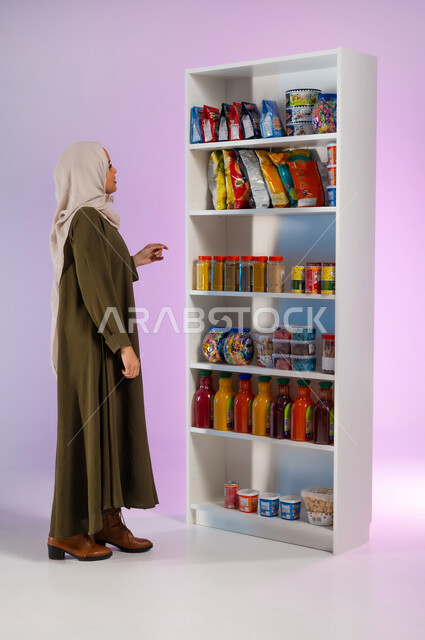 أصناف وأنواع من مستلزمات المستهلك من مواد غذائيه مختلفه في المراكز التجاريه, الاستفادة من العروض والتخفيضات الموسمية على السلع, بورتريه من الجانب لإمرأة عربية خلجية سعودية محجبة ترتدي العباءة تقوم بالتسوق والتبضع من السوبرماركت, صورة الجسم بالكامل, خلفية ملونة
