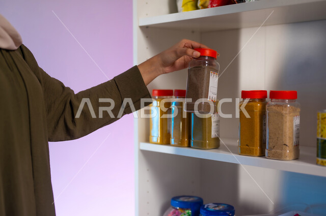 أنواع عديدة من البهارات في البقالة, عرض منتجات وتموينات محلية ومستوردة في المحلات التجارية, بورتريه مقرب من الجانب لسيدة عربية سعودية خليجية ترتدي العباءة الزيتية تقوم باختيار نوع من أنواع التوابل, متعة التسوق والشراء من السوبرماركت, خلفية ملونة