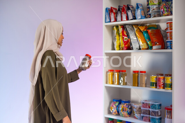 قراءة صلاحية المنتج ومكوناته, المواد الغذائية المختلفة والمنتجات المحلية في المراكز التجارية, السلع اللازمة لتلبية احتياجات المواطن السعودي, بورتريه من الجانب لشابة عربية خليجية سعودية ترتدي العباءة تمسك بيدها نوع من أنواع التوابل, خلفية ملونة