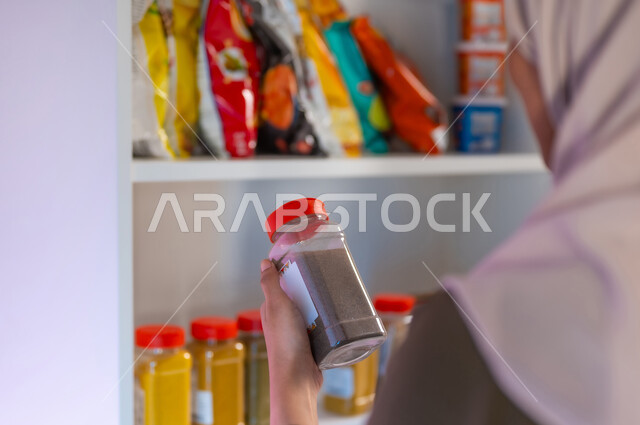 أنواع عديدة من البهارات في البقالة, صورة مقربة من الخلف لسيدة عربية سعودية خليجية محجبة تقوم باختيار نوع من أنواع التوابل, قراءة صلاحية المنتج ومحتوياته, قضاء وقت ممتع في الشراء والتسوق, عرض منتجات وتموينات محلية ومستوردة في المحلات التجارية