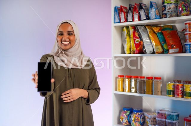 Purchasing essential foodstuffs through electronic applications, enjoying shopping in the supermarket, a close-up portrait of a smiling Saudi Gulf Arab woman wearing an olive abaya, displaying a mobile phone with a blank black screen, commercial markets to meet the needs of the Saudi citizen, white background