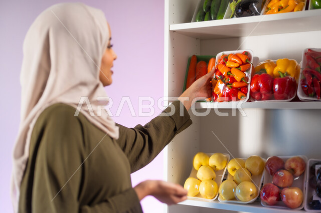 التبضع والشراء من قسم الخضراوات في السوبرماركت, المواد الغذائية على الرفوف في المحلات والمراكز التجارية, بورتريه مقرب من الجانب لإمرأة عربية خليجية سعودية محجبة ترتدي العباءة الزيتية تقوم بحمل صحن من الفلفل في البقالة, منتجات محلية وطنية سعودية, خلفية ملونة