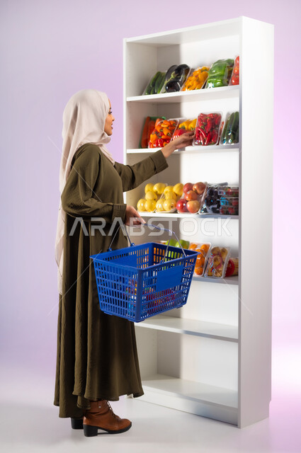 اقتناء أجود أنواع الخضار والفواكه من البقالة، شراء المواد الغذائية لتلبية احتياجات المواطن السعودي, بورتريه من الجانب لإمرأة عربية خليجية سعودية محجبة ترتدي العباءة الزيتية تمسك بيدها سلة التسوق في السوبرماركت, صورة الجسم بالكامل, خلفية ملونة