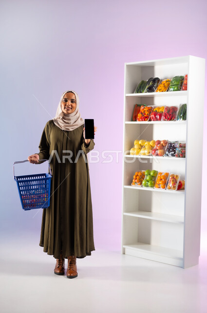 Mobile phone display with a blank black screen, buying vegetables and fruits from the grocery store, purchasing food items to meet the needs of the Saudi citizen, portrait of a smiling Saudi Arabian Gulf woman wearing an olive abaya holding a shopping basket in the supermarket in her hand, full body portrait, white background