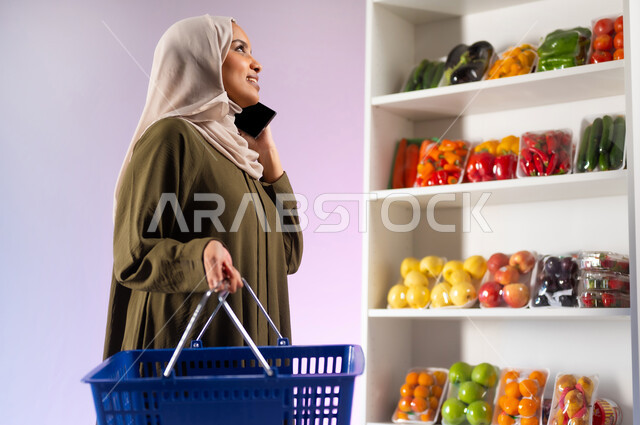 A commercial center to meet the needs of the Saudi consumer, shopping from the vegetable section in the supermarket, purchasing food items and supplies from the grocery store, a portrait from the side of a Saudi Arabian Gulf woman wearing an abaya, holding a shopping basket in her hand and making a voice call via mobile phone, white background