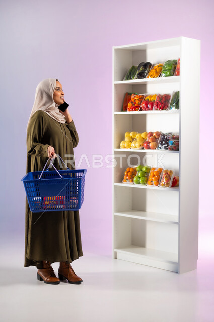 التبضع من قسم الخضراوات في السوبرماركت, شراء المواد والمستلزمات الغذائية من البقالة, بورتريه لشابة عربية خليجية سعودية ترتدي العباءة الملونة تمسك بيدها سلة التسوق وتقوم بإجراء مكالمة صوتية عبر الهاتف المحمول, صورة الجسم بالطول الكامل, خلفية ملونة