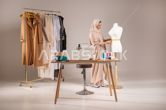 مهن و أعمال الخياطة و التصميم , تفصيل الملابس النسائية , بورتريه لمصممة أزياء عربية خليجية سعودية ترتدي الزي الكاجوال والحجاب تقف أمام المانيكان تأخذ القياسات بالشريط المتري , صورة للجسم بالطول الكامل , خلفية رمادية