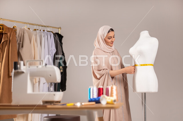 ابتكار تصاميم جديدة للألبسة النسائية , ممارسة مهنة الخياطة , بورتريه مقرب لمصممة أزياء عربية خليجية سعودية ترتدي الزي الكاجوال و الحجاب تأخذ القياسات بالشريط المتري , مهن و حرف يدوية , خلفية رمادية