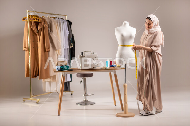 استخدام معدات و أدوات الخياطة , تصميم و تفصيل الملابس النسائية , بورتريه لمصممة أزياء عربية خليجية سعودية ترتدي الزي الكاجوال والحجاب تقف أمام المانيكان تأخذ القياسات بالشريط المتري , صورة للجسم بالطول الكامل , خلفية رمادية