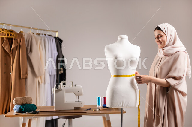 أدوات و معدات الخياطة , استخدام الشريط المتري لأخذ القياسات , بورتريه مقرب لمصممة أزياء عربية خليجية سعودية ترتدي الزي الكاجوال و الحجاب تقف أمام المانيكان , تصميم الألبسة النسائية  , خلفية رمادية