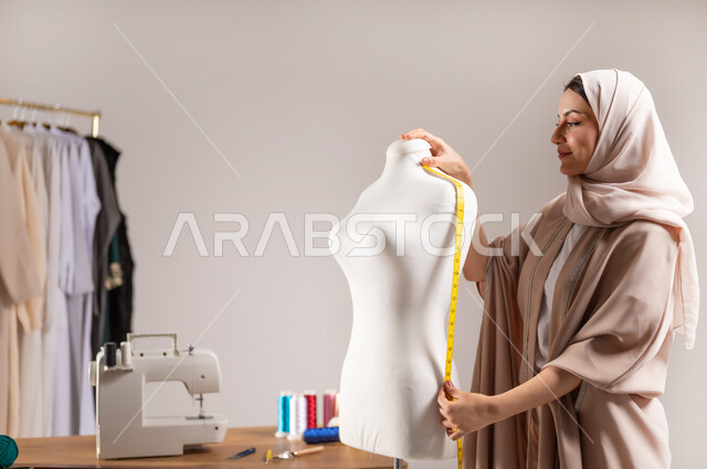 أخذ قياس الكتف بالشريط المتري , خياطة و تفصيل الملابس النسائية , بورتريه مقرب لمصممة أزياء عربية خليجية سعودية ترتدي الزي الكاجوال و الحجاب تقف أمام المانيكان , صناعة الألبسة في السعودية , خلفية رمادية