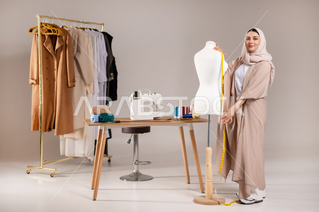 استخدام الشريط المتري لأخذ القياسات , خياطة و تفصيل الملابس النسائية , بورتريه لمصممة أزياء عربية خليجية سعودية ترتدي الزي الكاجوال والحجاب تقف أمام المانيكان , صورة للجسم بالطول الكامل , خلفية رمادية