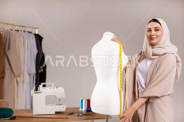 أحذ القياسات باستخدام الشريط المتري , النظر إلى الكاميرا بإيماءات السعادة و السرور , بورتريه مقرب لمصممة أزياء عربية خليجية سعودية ترتدي الزي الكاجوال و الحجاب تقف أمام المانيكان , ممارسة مهنة الخياطة , خلفية رمادية