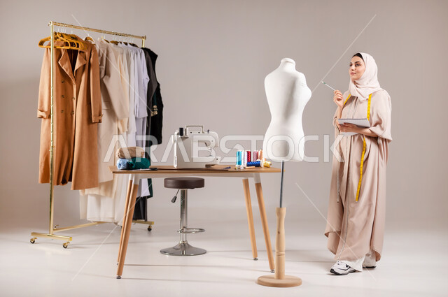 مهارات التخطيط و التصميم , مهن و حرف يدوية , بورتريه لمصممة أزياء عربية خليجية سعودية ترتدي الزي الكاجوال والحجاب تقف أمام المانيكان تمسك دفتر و قلم بيديها  , صورة للجسم بالطول الكامل , خلفية رمادية