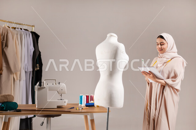 الموهبة و الإبداع في التصميم , ممارسة مهنة الخياطة , بورتريه مقرب لمصممة أزياء عربية خليجية سعودية ترتدي الزي الكاجوال و الحجاب تقف أمام المانيكان تمسك دفتر و قلم بيديها , صناعة الألبسة النسائية في السعودية , خلفية رمادية