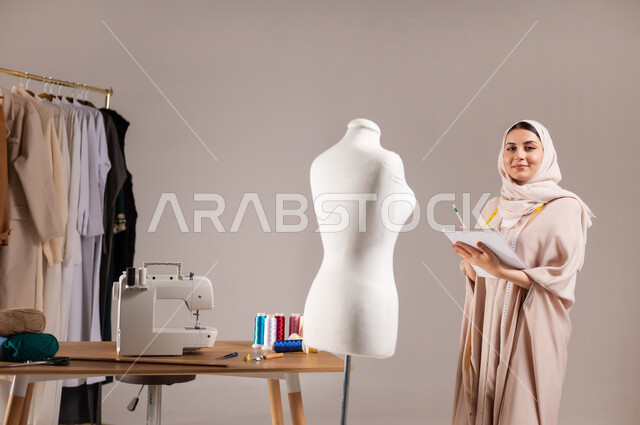 الإبداع في ابتكار موديلات جديدة , فنون الخياطة و التصميم , بورتريه مقرب لمصممة أزياء عربية خليجية سعودية ترتدي الزي الكاجوال و الحجاب تقف أمام المانيكان تمسك دفتر و قلم بيديها , النظر إلى الكاميرا بإيماءات السعادة و السرور , خلفية رمادية