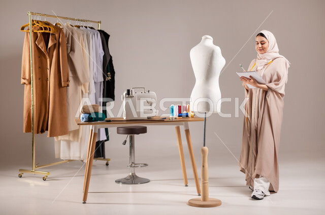 صناعة الأثواب النسائية السعودية , العمل في ورشة الخياطة , بورتريه لمصممة أزياء عربية خليجية سعودية ترتدي الزي الكاجوال والحجاب تقف أمام المانيكان تحمل دفتر و قلم بيدها , صورة للجسم بالطول الكامل , خلفية رمادية