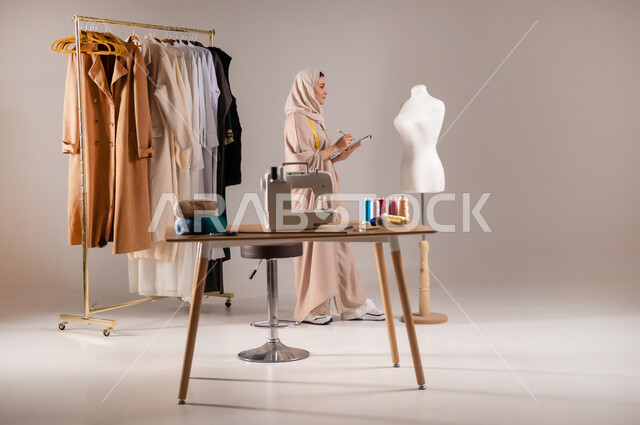 محل لتصميم الألبسة النسائية , ورشة خياطة في السعودية , بورتريه لمصممة أزياء عربية خليجية سعودية ترتدي الزي الكاجوال والحجاب , ابتكار موديلات جديدة , صورة للجسم بالطول الكامل , خلفية رمادية