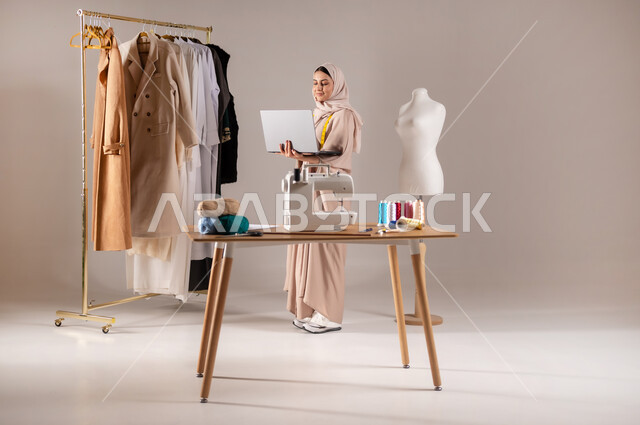 العمل في ورشة الخياطة , استخدام التقنيات الحديثة و المتطورة , بورتريه لمصممة أزياء عربية خليجية سعودية ترتدي الزي الكاجوال والحجاب تحمل جهاز الحاسوب بيديها , صورة للجسم بالطول الكامل , خلفية رمادية