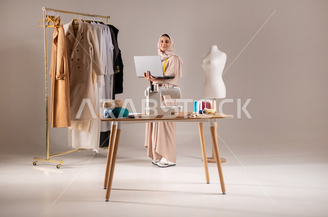 إنجاز المهام على الحاسب المحمول , ورشة عمل خياطة للألبسة النسائية , بورتريه لمصممة أزياء عربية خليجية سعودية ترتدي الزي الكاجوال والحجاب تحمل جهاز الحاسوب بيديها , صورة للجسم بالطول الكامل , خلفية رمادية