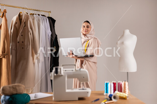 استخدام جهاز الحاسوب في ورشة العمل , إنجاز طلبات الزبائن , بورتريه مقرب لمصممة أزياء عربية خليجية سعودية ترتدي الزي الكاجوال و الحجاب , محل خياطة للألبسة النسائية , خلفية رمادية