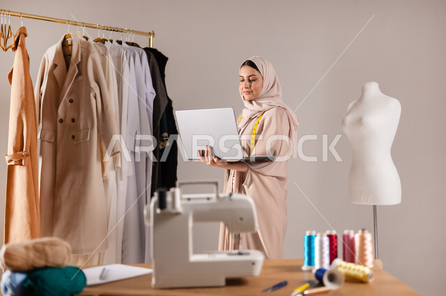 Using advanced technology at work, practicing tailoring, a close-up portrait of a Saudi Arabian Gulf fashion designer wearing casual clothes and a hijab, holding a computer in her hands, tailoring women’s clothing, gray background