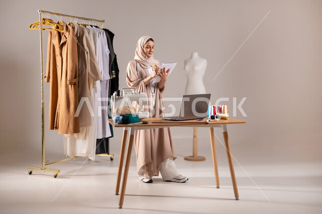إتقان رسم تصاميم الألبسة السعودية , خياطة و تفصيل الملابس النسائية , بورتريه لمصممة أزياء عربية خليجية سعودية ترتدي الزي الكاجوال والحجاب تمسك قلم و دفتر بيديها , صورة للجسم بالطول الكامل , خلفية رمادية