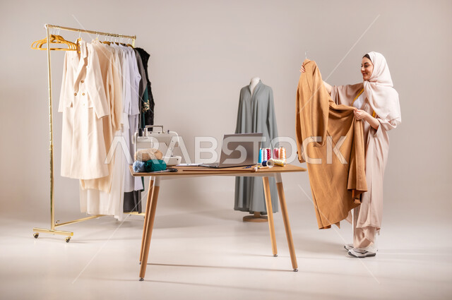 Fulfilling customer orders, practicing the sewing profession, portrait of a Saudi Arabian Gulf fashion designer wearing casual clothes and the hijab, holding a dress in her hands, women’s clothing design workshop, full-length body portrait, gray background