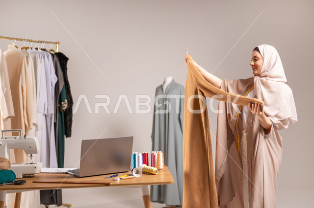 الاستمتاع بممارسة مهنة الخياطة , تفصيل الملابس النسائية , بورتريه مقرب لمصممة أزياء عربية خليجية سعودية ترتدي الزي الكاجوال و الحجاب تحمل ثوب بيديها , حرف و مهن يدوية , خلفية رمادية