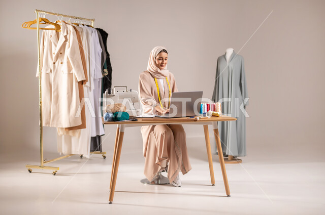 استخدام التكنولوجيا المتطورة في العمل , ممارسة مهنة الخياطة , بورتريه لمصممة أزياء عربية خليجية سعودية ترتدي الزي الكاجوال و الحجاب تقوم بإنجاز المهام على الحاسوب , ورشة تصميم أزياء , صورة للجسم بالطول الكامل , خلفية رمادية
