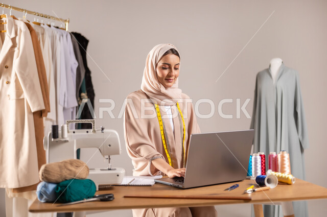 Using modern technologies at work, a sewing workshop for women’s clothes, a close-up portrait of a Saudi Arabian Gulf fashion designer wearing casual clothes and the hijab, using a laptop to complete tasks, fashion design and detailing, gray background