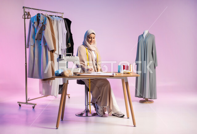 ابتكار تصاميم جديدة , العمل في ورشة خياطة , بورتريه لمصممة أزياء عربية خليجية سعودية ترتدي الزي الكاجوال و الحجاب تقوم بالرسم على الورق , صورة للجسم بالطول الكامل , خلفية ملونة