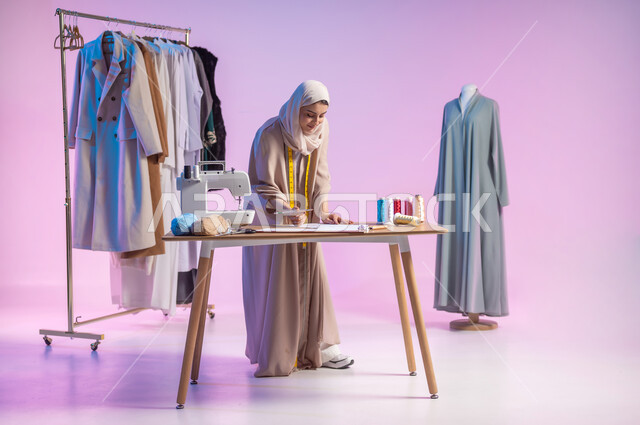 ابتكار و رسم تصاميم جديدة , ممارسة مهنة الخياطة , بورتريه لمصممة أزياء عربية خليجية سعودية ترتدي الزي الكاجوال و الحجاب تقوم بالرسم على الورق , صورة للجسم بالطول الكامل , خلفية ملونة