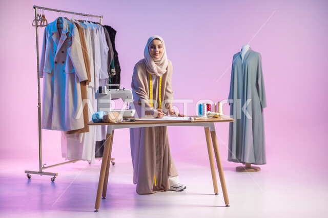 رسم التصاميم الجديدة , النظر إلى الكاميرا بإيماءات السعادة و السرور  , بورتريه لمصممة أزياء عربية خليجية سعودية ترتدي الزي الكاجوال و الحجاب تمسك القلم بيدها , العمل في ورشة خياطة , صورة للجسم بالطول الكامل , خلفية ملونة
