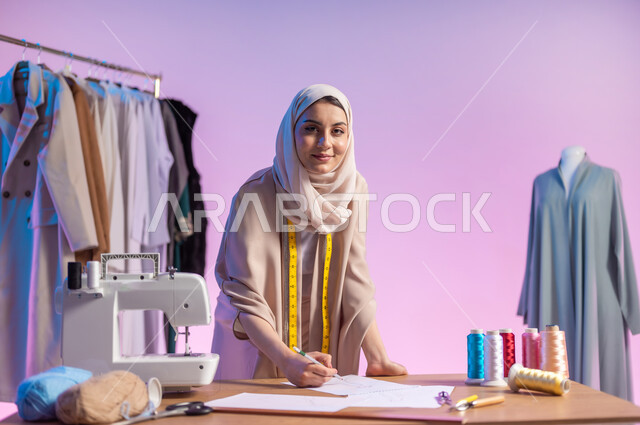 النظر إلى الكاميرا بإيماءات السعادة و السرور , العمل في ورشة خياطة , بورتريه مقرب لمصممة أزياء عربية خليجية سعودية ترتدي الزي الكاجوال و الحجاب تقوم برسم التصاميم على الورق , خلفية ملونة