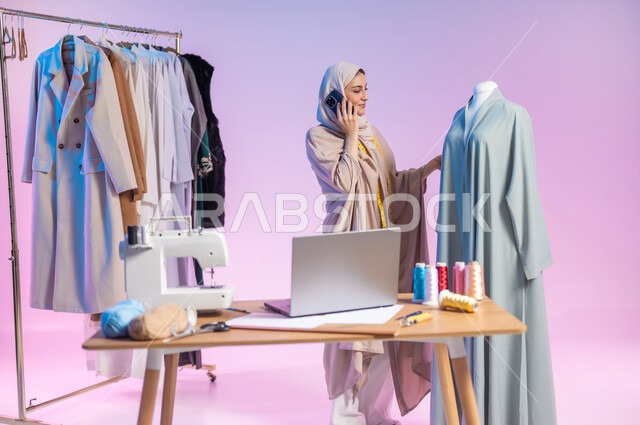 إجراء مكالمة هاتفية عبر الهاتف المحمول , العمل في ورشة الخياطة , بورتريه لمصممة أزياء عربية خليجية سعودية ترتدي الزي الكاجوال و الحجاب تقف أمام المانيكان , مهن و حرف يدوية , صورة للجسم بالطول الكامل , خلفية ملونة