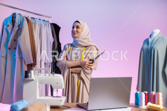 ممارسة مهنة تصميم الأزياء , ورشة خياطة ملابس نسائية , بورتريه مقرب لمصممة أزياء عربية خليجية سعودية ترتدي الزي الكاجوال و الحجاب تمسك المقص بيدها  , إيماءات الفرح و السرور , خلفية ملونة