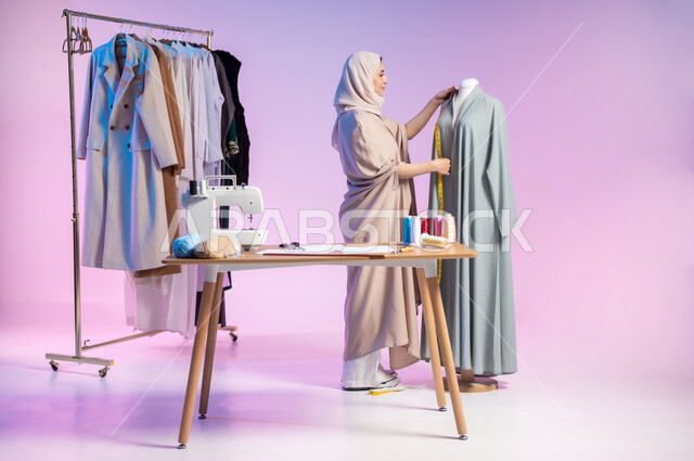 استخدام المتر لقياس القماش , ممارسة مهنة الخياطة , بورتريه لمصممة أزياء عربية خليجية سعودية ترتدي الزي الكاجوال و الحجاب تقوم بأخذ القياسات المناسبة , صورة للجسم بالطول الكامل , خلفية ملونة