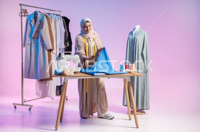 تصميم الملابس النسائية , العمل في ورشة الخياطة , بورتريه لمصممة أزياء عربية خليجية سعودية ترتدي الزي الكاجوال و الحجاب تقوم بقص قطعة القماش , استخدام الأدوات المناسبة لتفصيل الثياب , صورة للجسم بالطول الكامل , خلفية ملونة