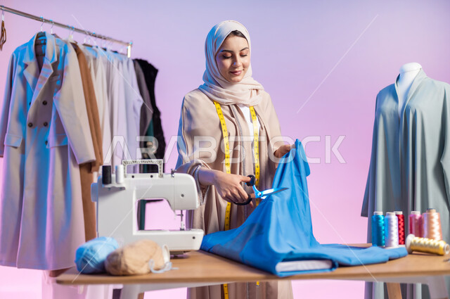 العمل في ورشة الخياطة , تصميم الملابس النسائية , بورتريه مقرب لمصممة أزياء عربية خليجية سعودية ترتدي الزي الكاجوال و الحجاب تقوم بقص قطعة القماش , استخدام الأدوات المناسبة تفصيل الثياب , خلفية ملونة
