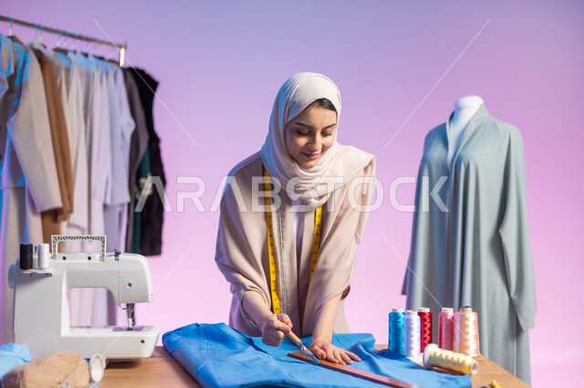 Using appropriate tools to detail clothes, fashion design professions and works, close-up portrait of a Saudi Arabian Gulf fashion designer wearing a casual outfit and hijab, holding a scalpel and ruler, colored background