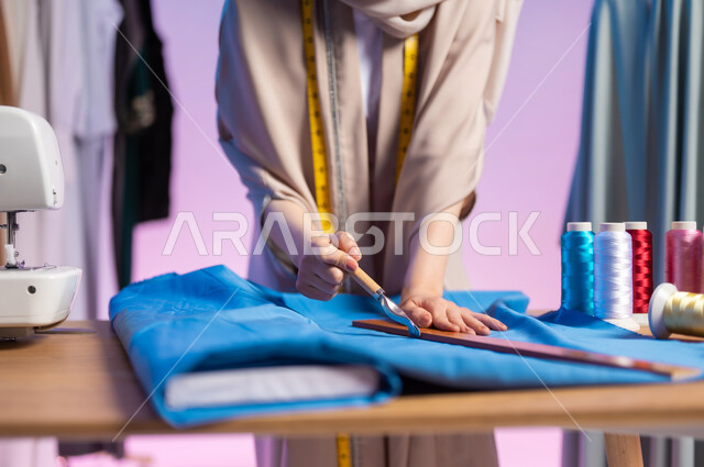 مهن و أعمال تصميم الأزياء , استخدام الأدوات المناسبة لتفصيل الملابس , بورتريه مقرب لمصممة أزياء عربية خليجية سعودية ترتدي الزي الكاجوال و الحجاب تمسك المشرط و المسطرة  , خلفية ملونة