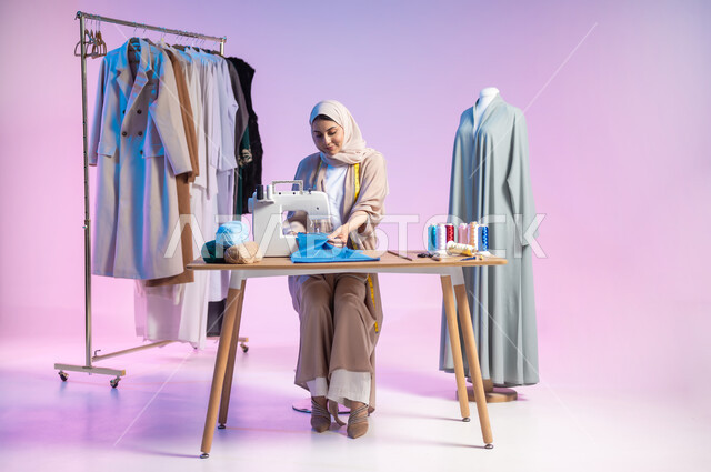 تفصيل الثياب باستخدام ماكينة الخياطة , مهارة تصميم الملابس , بورتريه لمصممة أزياء عربية خليجية سعودية ترتدي الزي الكاجوال و الحجاب , صورة للجسم بالطول الكامل , حرف و مهن يدوية في السعودية , خلفية ملونة